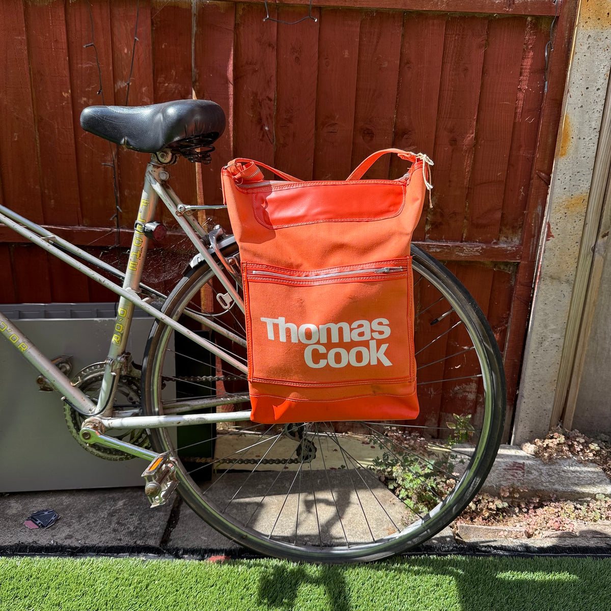An orange Thomas Cook bicycle pannier bag hanging from the rear rack of a silver bicycle, set against a wooden fence and outdoor ground. upcycled goodordering bag 