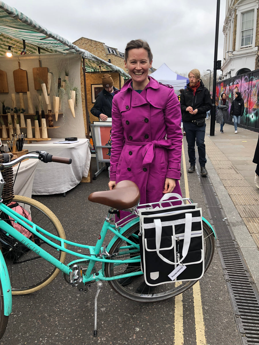 A woman in a bright magenta coat stands next to a turquoise city bicycle with a black front basket on a street. The photo is rotated sideways, and market stalls and pedestrians are visible in the background.